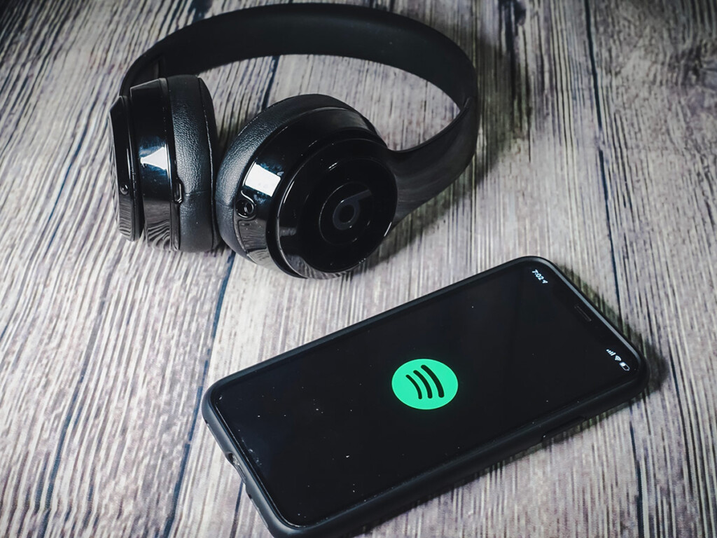 Sitting on a wooden surface, a Spotify app is open on a cellphone beside a pair of headphones.