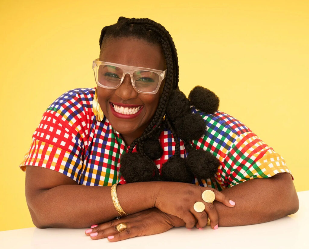 Nana aba Duncan dressed in a colourful outfit, sits smiling, arms crossed, against a yellow background.