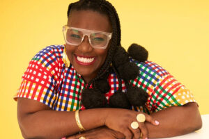 Nana aba Duncan dressed in a colourful outfit, sits smiling, arms crossed, against a yellow background.