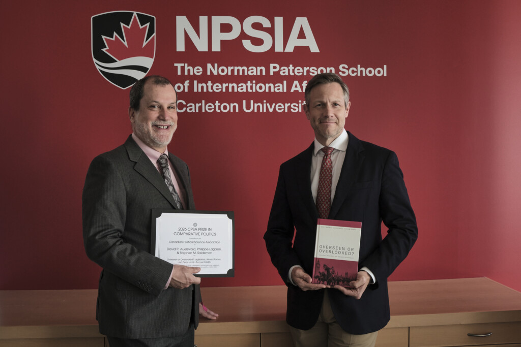 Philippe Lagassé and Stephen Saideman pose in front of a large NPSIA logo on a red wall
