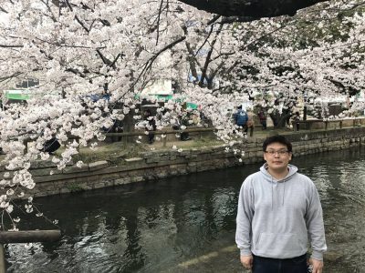 Ken Yoshida standing in front of a river and cherry blossoms