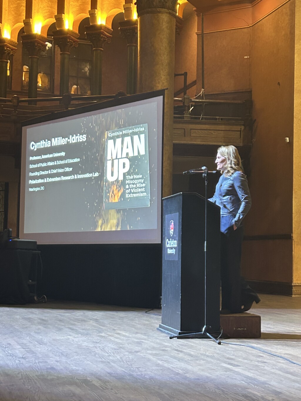 Cynthia Miller-Idriss standing at a podium next to a large screen presentation
