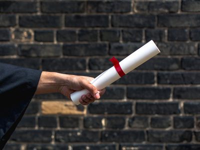 student holding out diploma in front of brick wall
