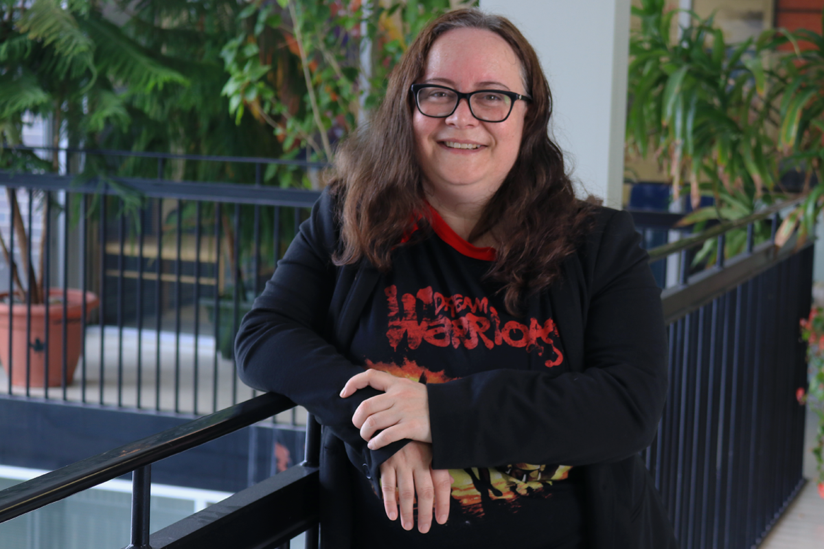 Prof. Laura Hall stands against a black railing, Laura has long brown hair and is wearing a black blazer over a black and red graphic tshirt. 