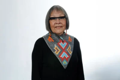 Indigenous women with short grey hair and glasses wearing Indigenous scarf and black sweater. 