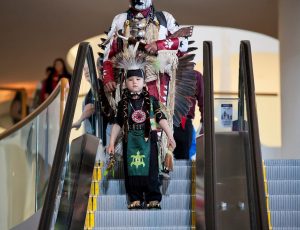 a young boy is walking down an escalator in traditional Indigenous clothing.