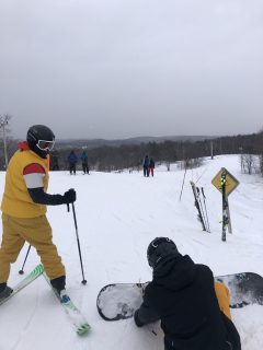 Two students at skill hill. One student on standing on skis, second student sitting on ground with snowboard.