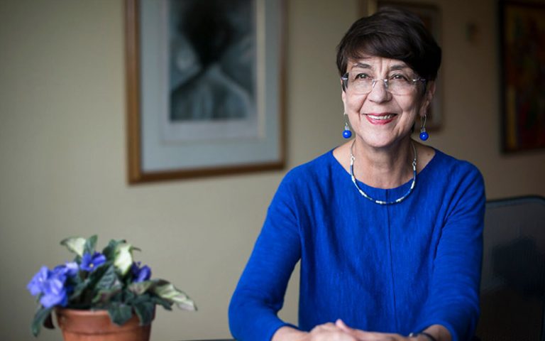 Cristina is a middle-aged woman Latinx woman with glasses, wearing a a vivid blue top and earrings of the same blue.