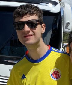 A young man with dark curly hair, wearing sunglasses and a Colombian football shirt