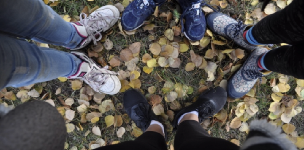 A photo of four people's pairs of shoes as they stand on the leaf-strewn ground.