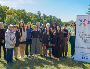 A group of people stand close together outside with a blue sky and green trees behind them. On the right, a white banner stands titled "LERRN, Local Engagement Refugee Research Network."