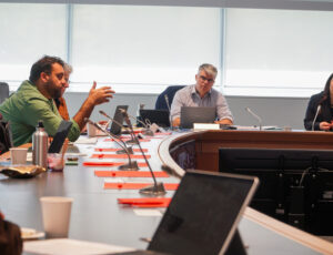 Two men and one woman sit around a horseshoe shaped table lined with red books and microphones. The man on the left has his hand out like he is making a point while he speaks. The others look at him as he speaks.