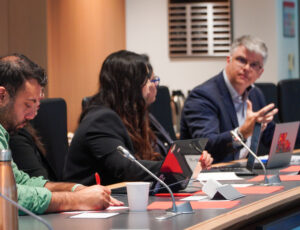 Three people sit in a row along a table. On the left, a man in a green shirt writes notes, in the middle a woman wearing black leans in to listen to the man on the right who looks back at her as he speaks.