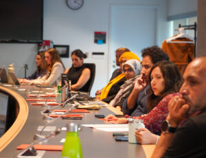 A group of people are seated. Some take notes others look across the room to the unpictured other side of the table.