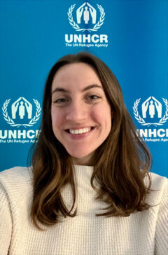 A head-and-shoulders portrait of Erika Ehrenberg with long, wavy brown hair and a friendly smile. She is wearing a white, textured knit sweater and a small nose stud. The background is a solid blue wall featuring three white UNHCR (The UN Refugee Agency) logos.
