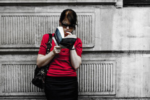 Woman reading a book while leaning on a wall.