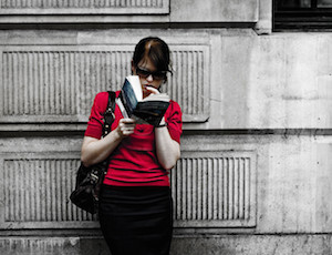 Woman reading a book while leaning on a wall.