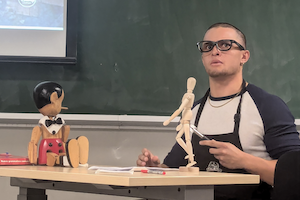 Student in glasses looks up. There is a wooden artist model on the table and Pinocchio puppet
