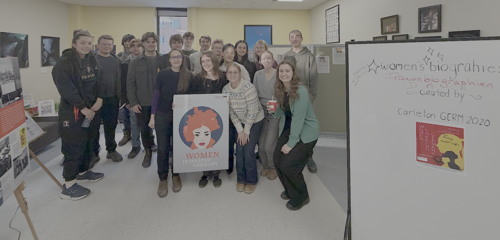 Group of German students pose around a poster that shows woman with bright red hair & board that reads "Women's Biographies"