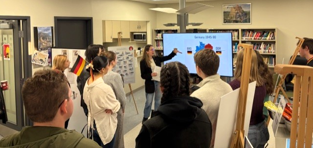 Group of students gathered to listen to rep from Embassy of Germany. PowerPoint shown on smart TV.