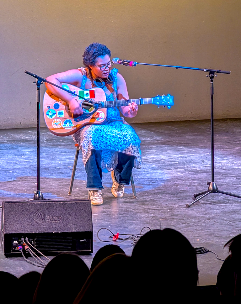 Madie plays a guitar covered with stickers including the Mexican flag.  She is seated in a chair.