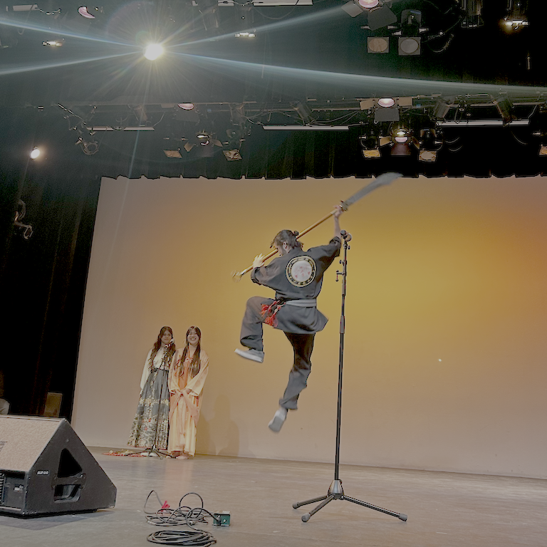 Mo leaps in the air with a staff while Natharie and Amie sing from their shared microphone.