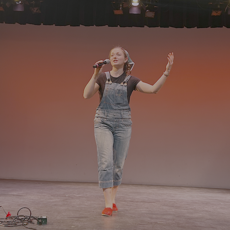 Chloé dressed in denim overalls, a bandana, and red shoes raises her hand urging people to join in.