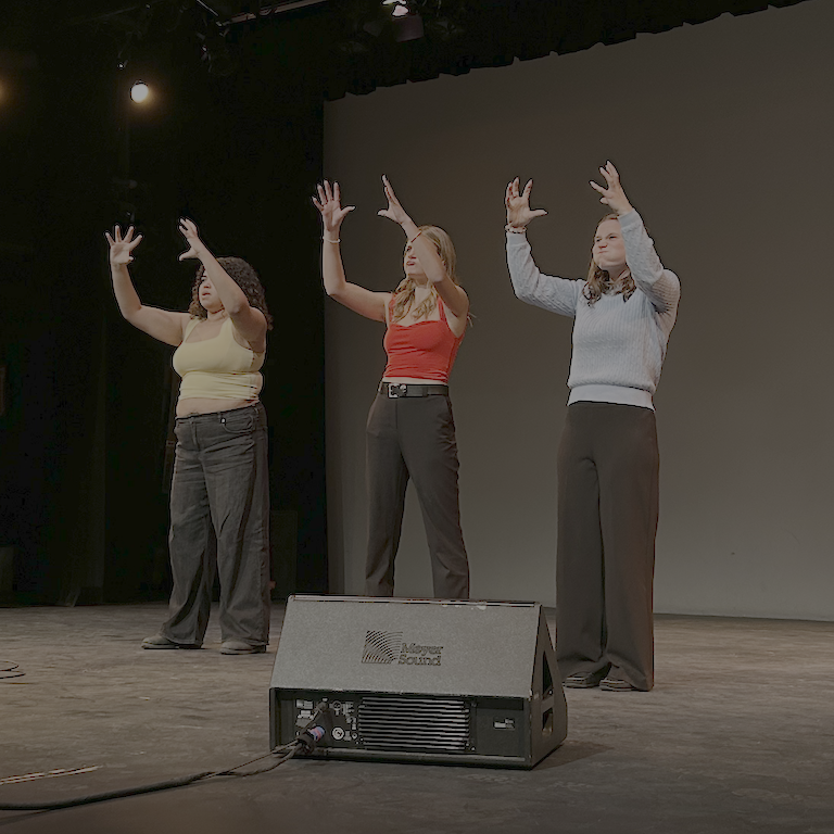 Three students hold their hands up to the sky as flowers, one in a yellow top, one red, and one blue.