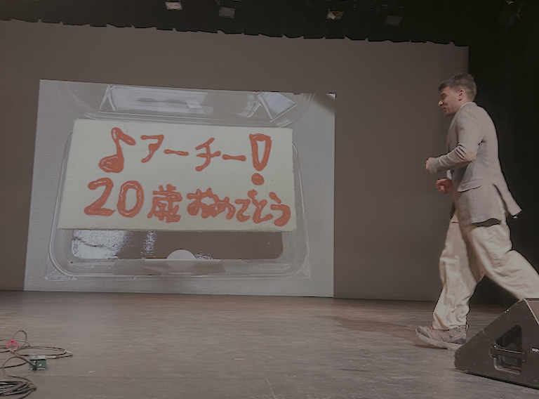 Archie dressed in a suit walks across the stage.  Behind him there is a birthday cake wishing him happy birthday in Japanese.