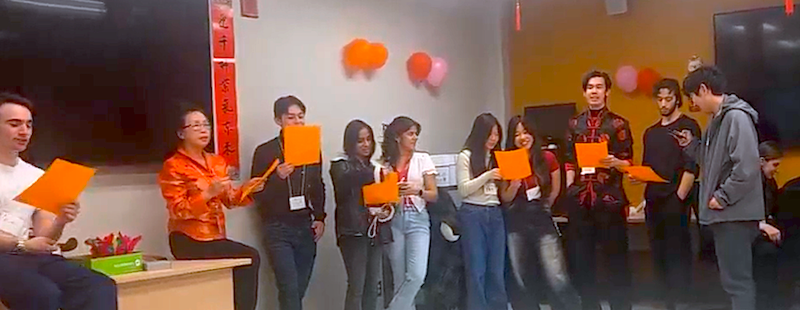 Group of students and instructor singing.  All are holding orange sheets of paper.