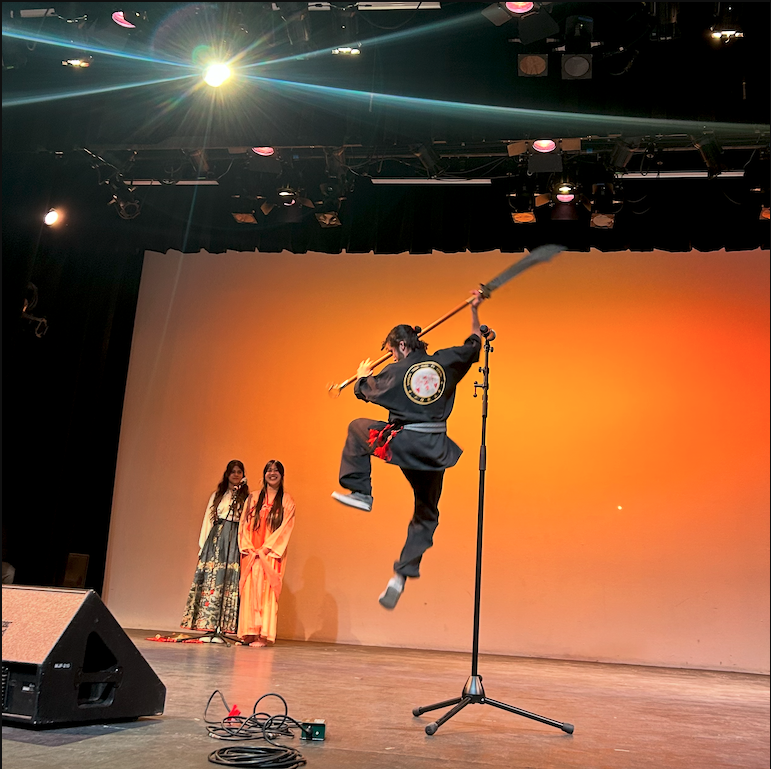 Mo leaps in the air with a staff while Natharie and Amie sing from their shared microphone.