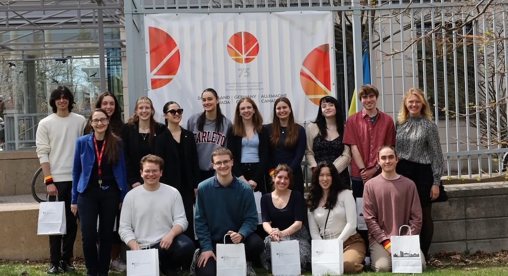 Students holding gift bags in front of Germany at 75 poster, mounted on a fence. Instructor & Embassy rep also in photo.