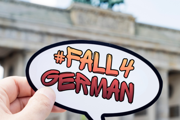 Hand holding a sticker that says #Fall4German. In the background, we see a blurry Brandenburg Gate.