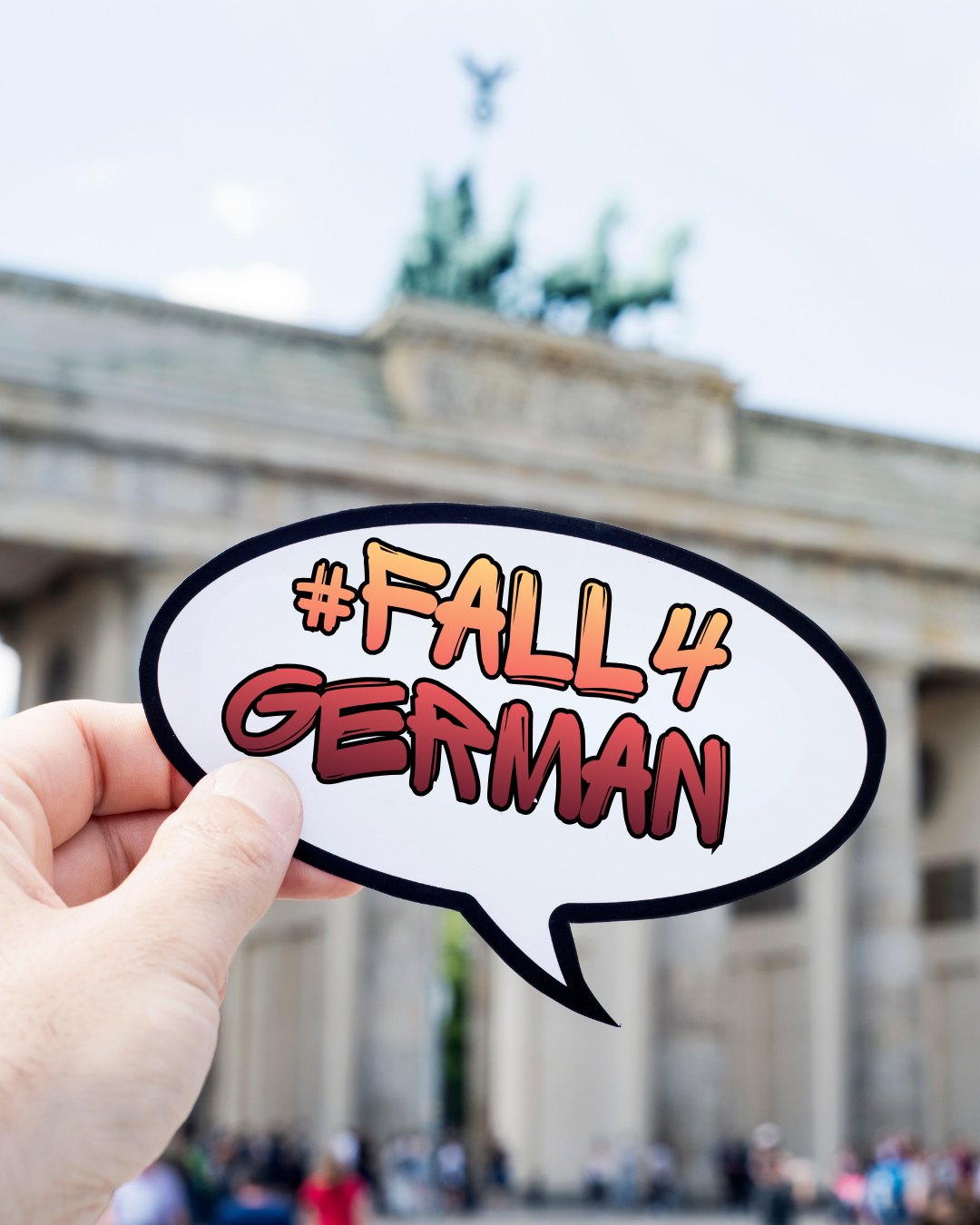 Hand holding a sticker that says #Fall4German. In the background, we see a blurry Brandenburg Gate.