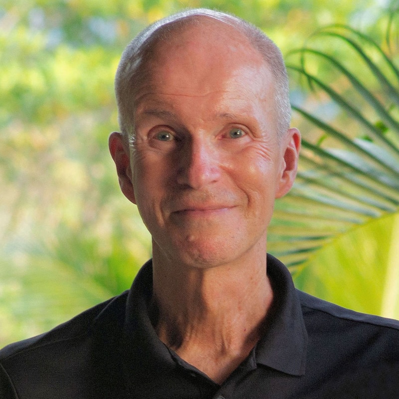 With palm leaves behind his head, a man in a dark shirt smiles at the camera.