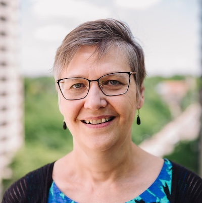 A woman (Cathy Barr) with earrings, glasses and short hair smiles at the camera.