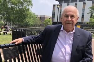 A man with a mauve dress shirt and suit jacket and slacks sits on a metal park bench with a recognition plaque bearing his name, Keith Sjogren.