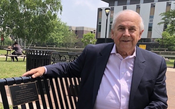 A man with a mauve dress shirt and suit jacket and slacks sits on a metal park bench with a recognition plaque bearing his name, Keith Sjogren.