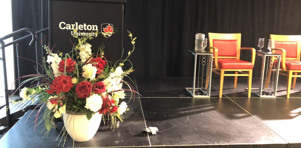 A huge bouquet of flowers sits in front of a black podium emblazoned with the Carleton University logo. Beside the podium are two empty chairs.