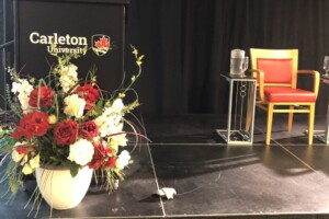 A huge bouquet of flowers sits in front of a black podium emblazoned with the Carleton University logo. Beside the podium are two empty chairs.