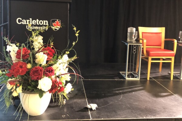 A huge bouquet of flowers sits in front of a black podium emblazoned with the Carleton University logo. Beside the podium are two empty chairs.