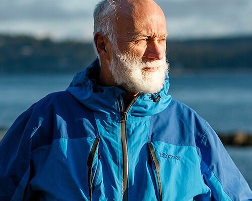 On a rugged beach in BC, a man, Eric, with a raincoat and arms on his hips stares into the distance to his left.