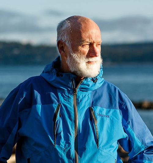 On a rocky beach in BC, a man, Eric, with a raincoat and arms on his hips stares into the distance to his left.