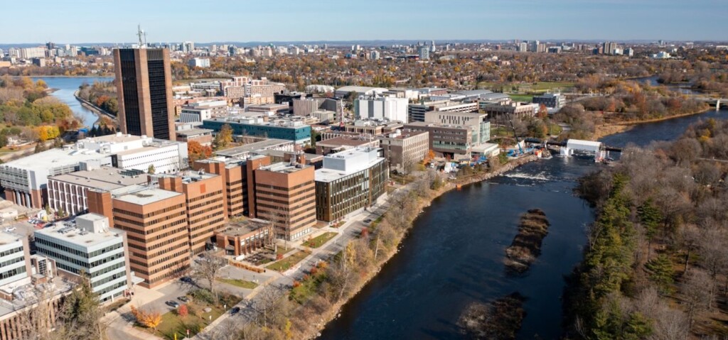 A long-range drone view of Carleton University shows the entire campus bordered by the Rideau River and Rideau Canal, with Ottawa spreading to the horizon in the background.