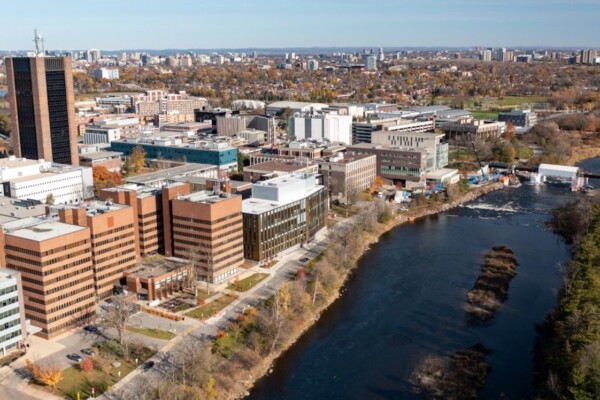 A long-range drone view of Carleton University shows the entire campus bordered by the Rideau River and Rideau Canal, with Ottawa spreading to the horizon in the background.