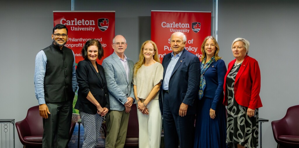 Seven people stand in a line in front of two Carleton University banners. They're smiling at the camera.