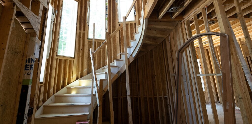 Inside of a new house being constructed, a beautiful wooden staircase curls to the second floor surrounded by cascading windows and half-built wooden walls.