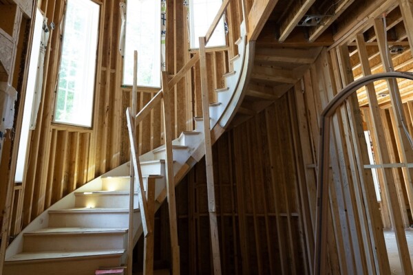Inside of a new house being constructed, a beautiful wooden staircase curls to the second floor surrounded by cascading windows and half-built wooden walls.