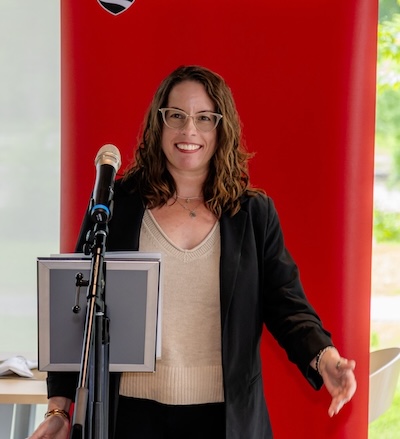 A woman (Jamie) speaks at a podium with Carleton's red banner behind her.