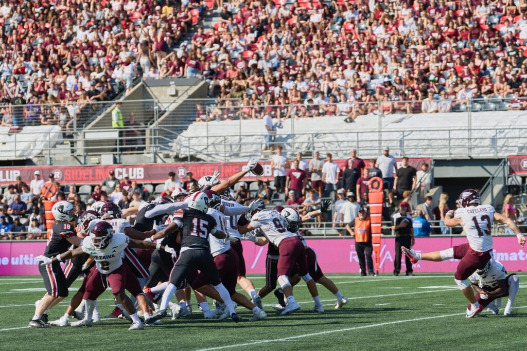 uOttawa Gee-Gees kicker attempts a field goal while Carleton Ravens defenders leap to block during an intense play.